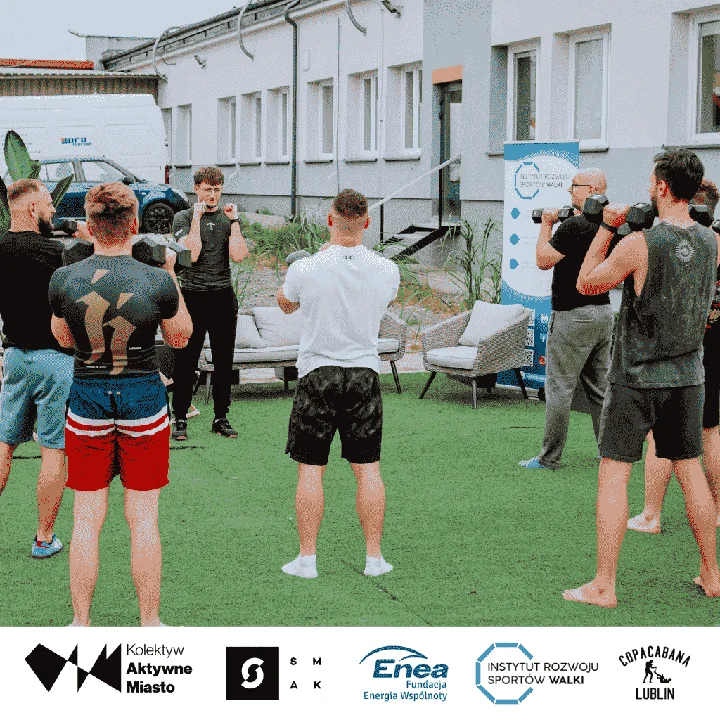 Grupa osób w sportowych strojach trenuje na sztucznej trawie na parkingu przed budynkiem. W tle widoczny trener, donice z roślinami, meble ogrodowe i leżaki tworzące zieloną strefę.