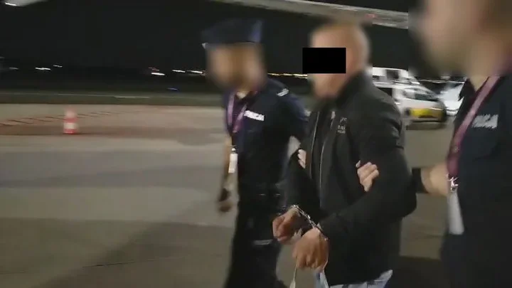 Mężczyzna w kajdankach jest prowadzony przez dwóch funkcjonariuszy na lotniskowym pasie nocą.