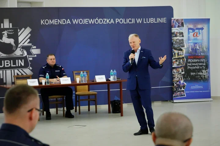 Mężczyzna w garniturze przemawia przez mikrofon podczas spotkania w siedzibie Komendy Wojewódzkiej Policji w Lublinie, przy stole siedzi policjant w mundurze.