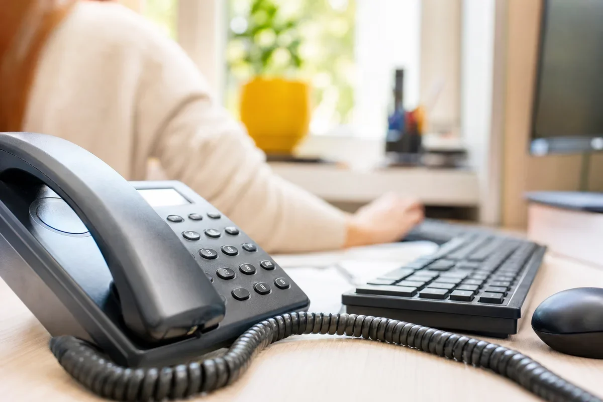 Biurko z telefonem stacjonarnym, klawiaturą i myszką, przy którym siedzi osoba pracująca przy komputerze.