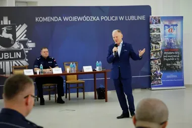 Mężczyzna w garniturze przemawia przez mikrofon podczas spotkania w siedzibie Komendy Wojewódzkiej Policji w Lublinie, przy stole siedzi policjant w mundurze.