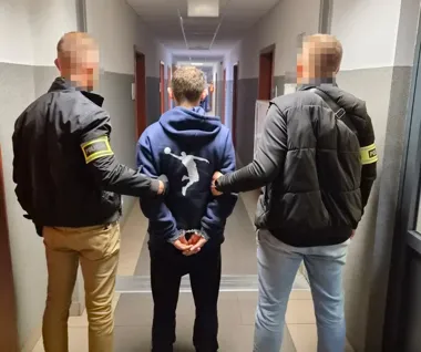 Zatrzymany mężczyzna w kajdankach prowadzony jest przez dwóch policjantów korytarzem budynku.