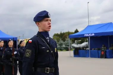 Młody mężczyzna w granatowym mundurze policyjnym z beretem stoi na baczność na placu, w tle namiot z napisem POLICJA i inne osoby w mundurach.