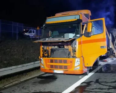 Pomarańczowa ciężarówka z uszkodzoną przednią częścią stoi nocą na poboczu drogi, z otwartą maską i drzwiami kabiny, w tle widać pojazd służbowy za ogrodzeniem.