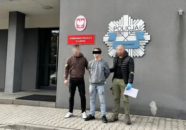 Zatrzymany mężczyzna w szarym swetrze stoi między dwoma funkcjonariuszami przed budynkiem Komisariatu Policji V w Lublinie.