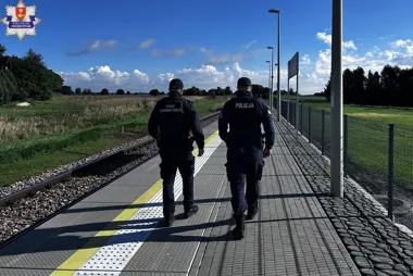 Funkcjonariusz policji i strażnik ochrony kolei patrolują peron kolejowy na otwartym terenie w słoneczny dzień.