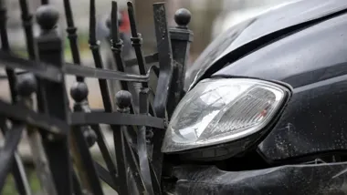 Uszkodzony czarny samochód osobowy zgnieciony przy czarnym metalowym ogrodzeniu.