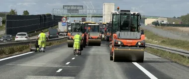 Robotnicy drogowi w odblaskowych kombinezonach walcują świeżo położoną nawierzchnię asfaltową na wielopasmowej drodze, przy ruchu pojazdów na sąsiednim pasie.